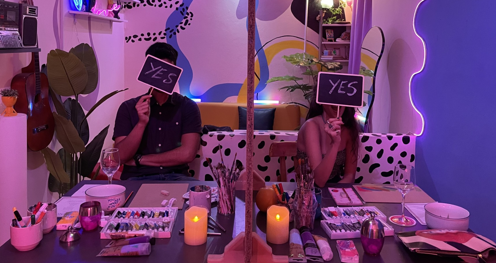 Two people on a blind date at the ICONIK studio, each holding up a YES sign, with paints and candles on the table between them